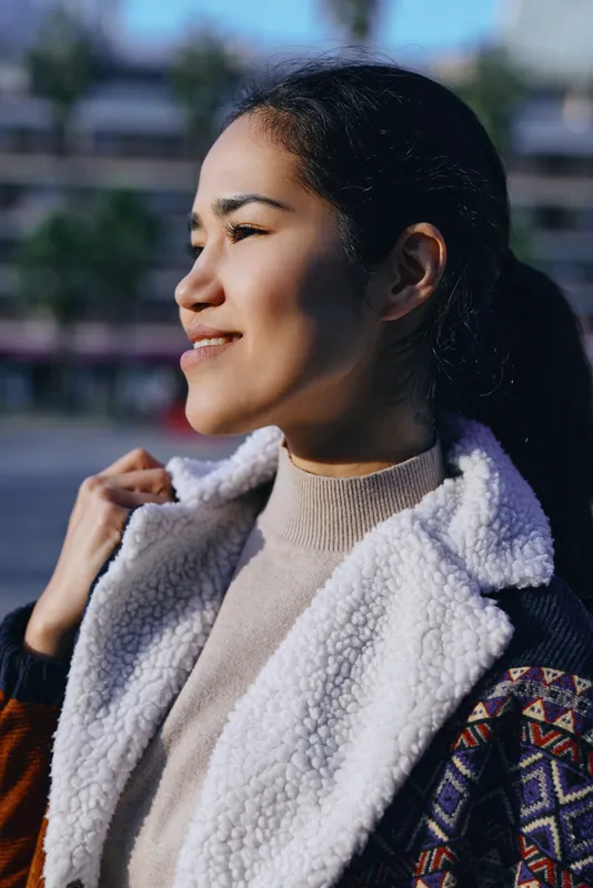 Smiling woman in a shearling coat outdoors, sunlight on her face, casual streetwear mood, candid lifestyle shot focused on fashion and everyday moments