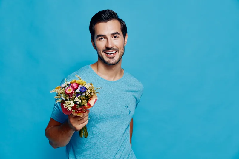 Smiling young man holding a bouquet of flowers, wearing a casual blue t-shirt, against a vibrant turquoise background, showcasing happiness and warmth.