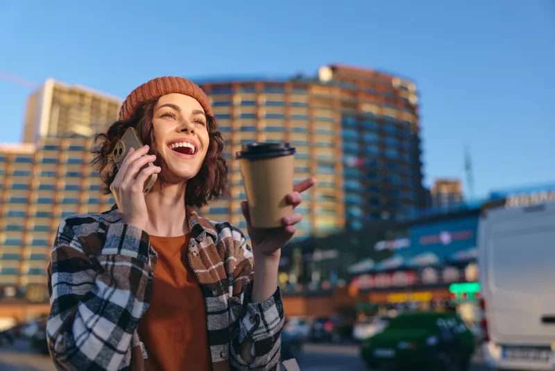 Woman with coffee and phone smiling in city street, beanie and jacket, urban sunlight and golden hour glow create candid lifestyle scene of authenticity and mindful living.