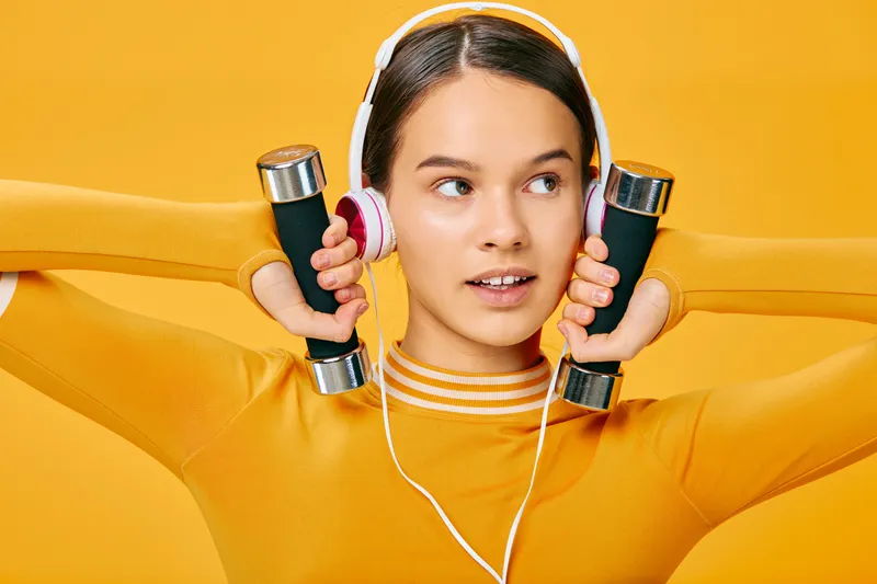Young woman in bright orange athletic wear lifts dumbbells resting on shoulders, wearing headphones against a vivid yellow backdrop for energetic fitness and workout motivation.