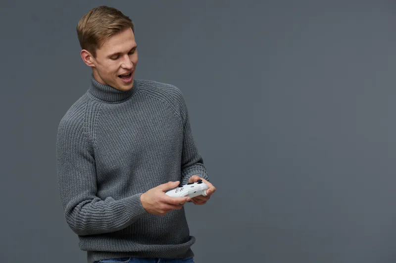 Young caucasian man wearing gray turtleneck sweater holding gaming controller with a smile, enjoying leisure time, isolated on plain gray background studio portrait. People lifestyle concept with posi