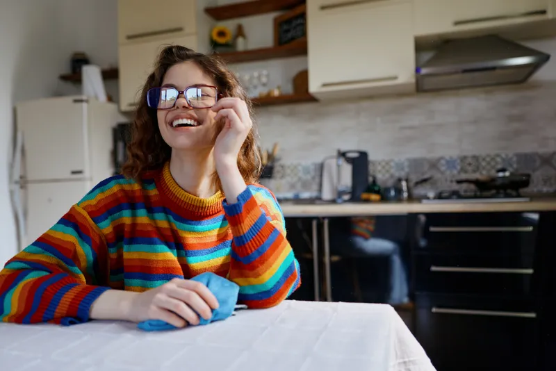 Young woman in a bright striped sweater sits at a kitchen table in a modern home, smiling warmly as she adjusts her glasses and enjoys a candid moment.