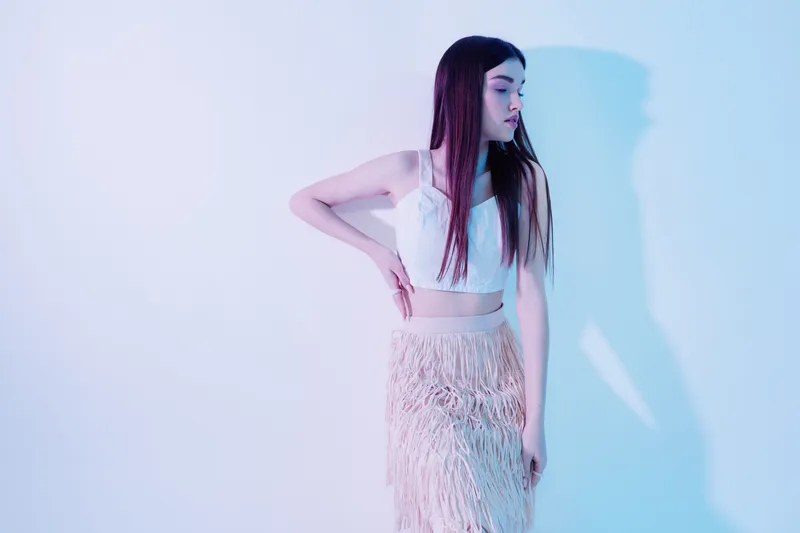 Young woman in white crop top and fringed skirt poses against light background. Her long hair is straight and she looks to the side. Concept of beauty fashion and style.