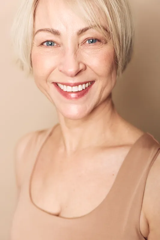 Smiling middle aged woman with short blonde hair and clear blue eyes wears beige top. Portrait on soft beige background with natural light. Concept for happiness, beauty, and confidence.