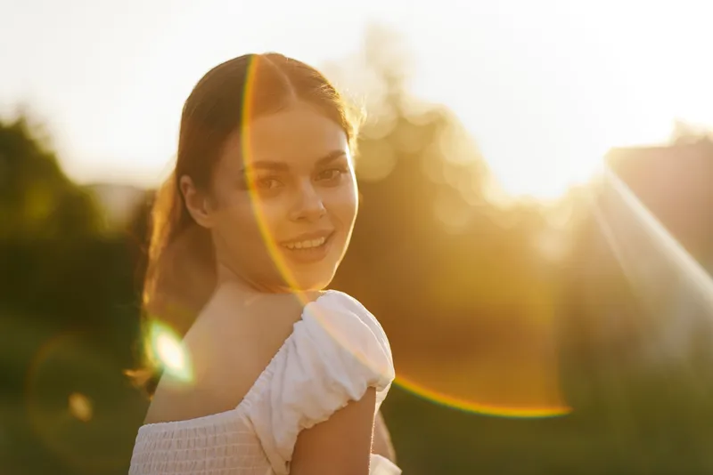 Young woman smiling in sunny outdoor setting, wearing white dress, exuding joy and positivity, surrounded by nature during golden hour