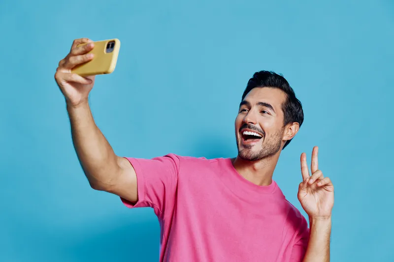Smiling young man wearing pink t-shirt takes selfie with smartphone against vibrant blue background. Cheerful, lifestyle concept, joy of capturing moments.