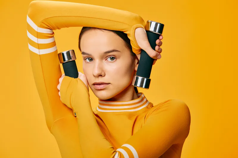 Woman in an athletic orange top lifts dumbbells in a bright studio, showcasing fitness, sport and workout focus with a strong pose and determined expression.