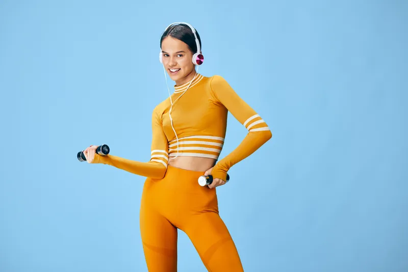 Woman in bright orange activewear poses with dumbbells against a blue studio backdrop, wearing headphones. Energetic fitness scene emphasizing sport, workout, exercise, and strength training in a