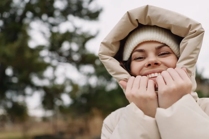Winter outdoor portrait of a smiling woman in a hooded coat, beanie and warm hands, cozy mood in a chilly natural setting with trees