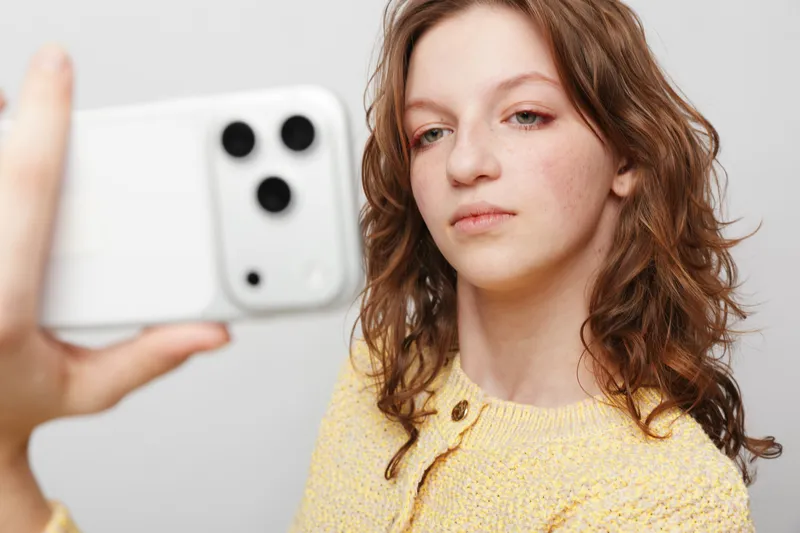 Young woman with curly hair wearing yellow sweater takes a selfie with a white smartphone on a light background. Concept of casual lifestyle, technology, and self-portrait photography.
