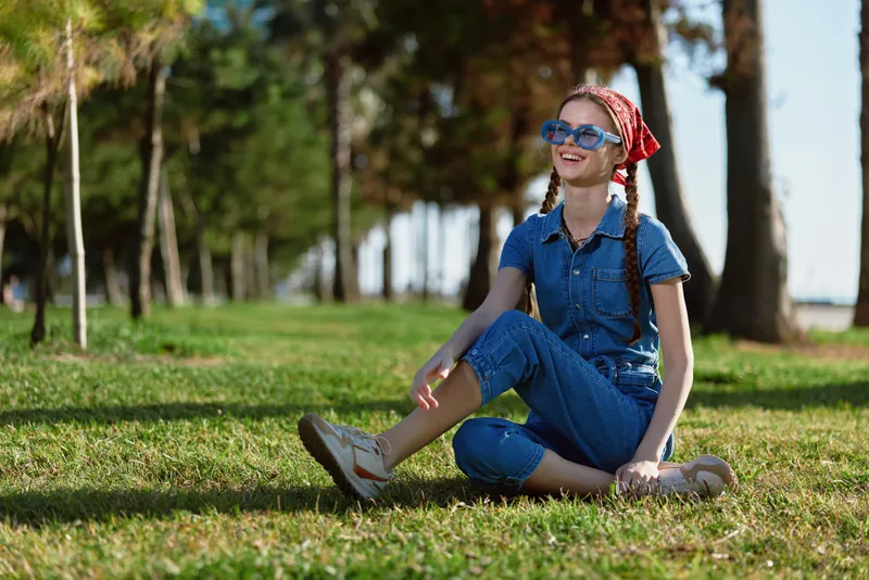 Lifestyle woman in modern boho-western denim street style sits on grass with sunglasses and bandana, bright film color capturing joyful outdoor moment in park.