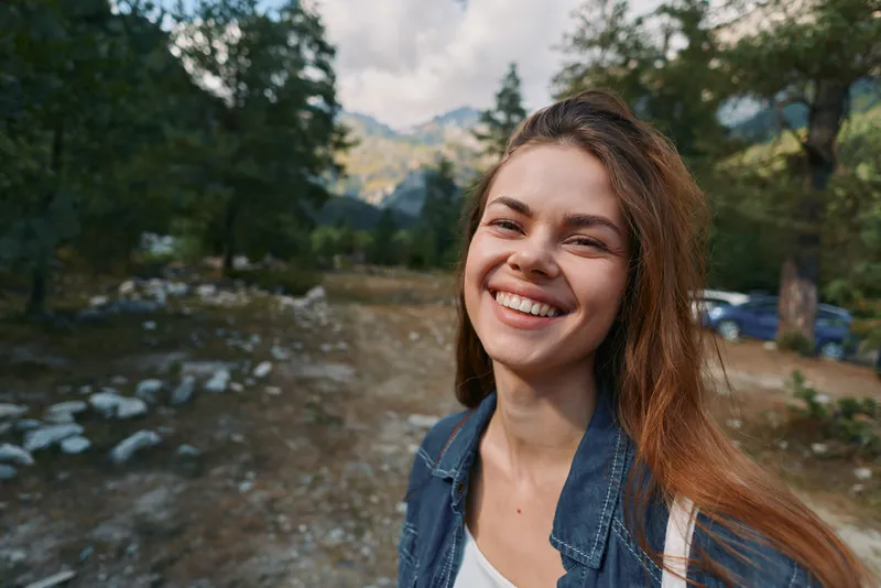 A cheerful woman stands outdoors in a natural setting, smiling warmly as sunlight glints on her denim jacket. She appears relaxed, confident, and ready for exploration.