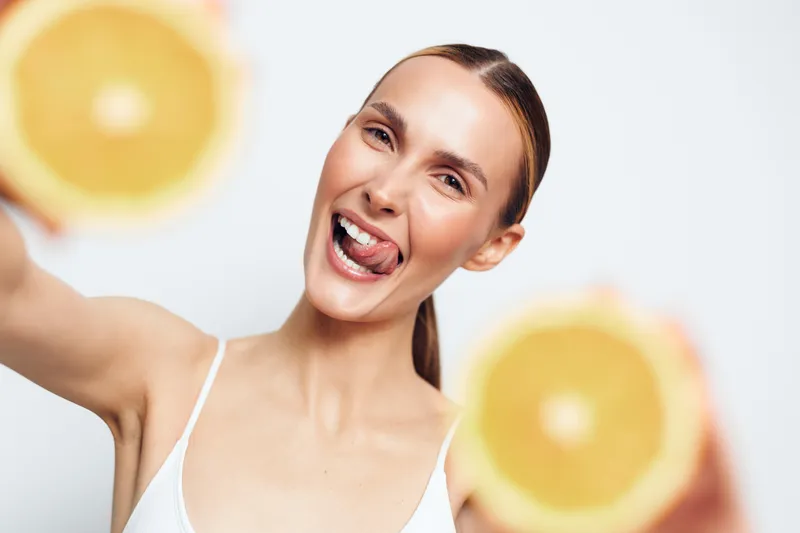 A young woman in a white tank top smiles brightly in a bright studio, reaching toward the camera with oranges in the foreground, capturing energy and freshness.