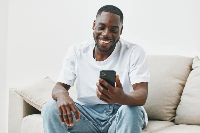 Happy young African American man relaxing on a couch while using a smartphone, dressed in a casual white t shirt, showcasing joy and comfort in a modern living space