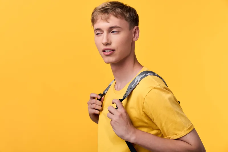 Male student wearing a yellow t shirt and backpack against a bright backdrop, ready for study and exploration, showcasing casual style, energy and confidence in a vivid studio scene.