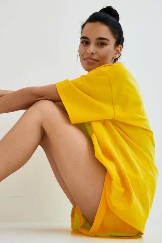 smiling woman in bright yellow oversized t-shirt sitting indoors, casual pose with natural makeup and bun hairstyle, soft lighting, minimalistic background, relaxed mood