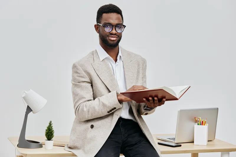 Professional young man with glasses, curly hair and dress shirt, standing in modern office, holding open book, ready to read or present, behind him a workspace with laptop, pen holder, paper notes and