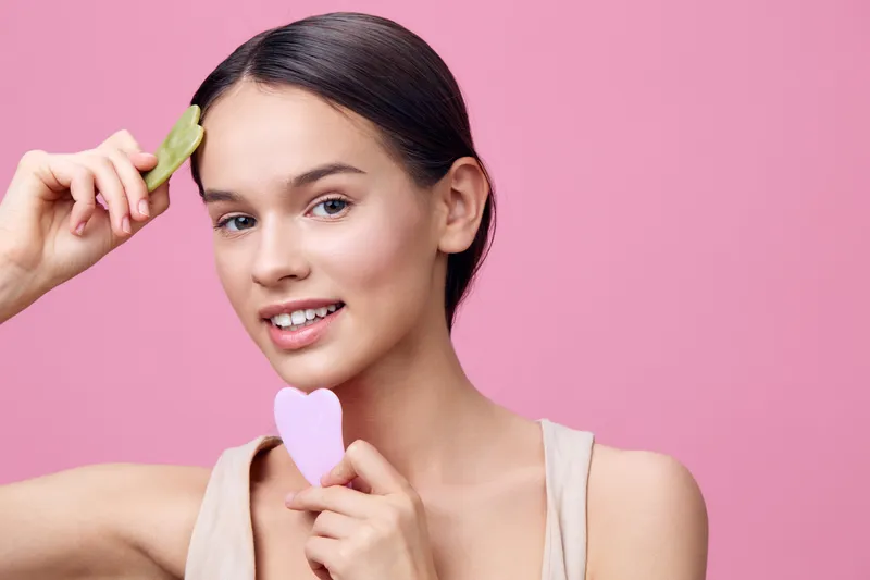 Young woman with a gua sha tool and heart shaped sponge in a skincare routine against a pink background, representing self care and beauty