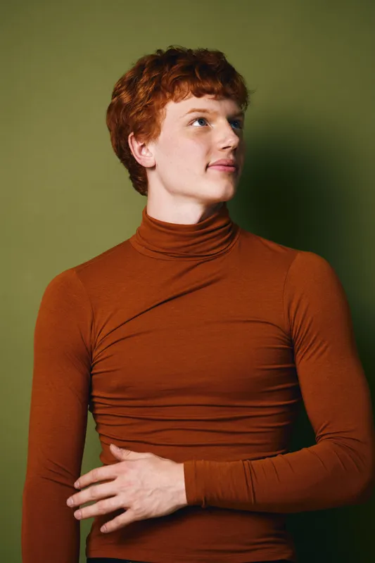 Portrait of young man with red hair wearing brown turtleneck sweater looking sideways against green background. Casual style with natural light and thoughtful expression.