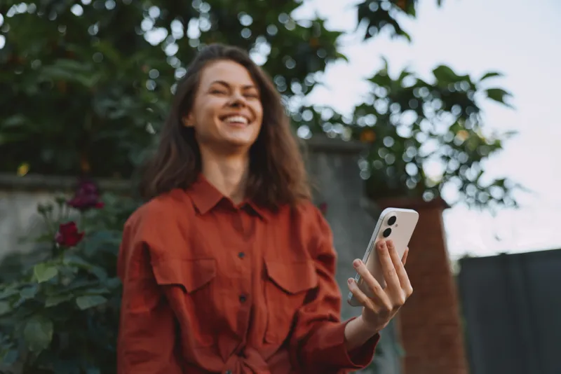Woman lifestyle portrait outdoors, smiling woman holding smartphone in casual red shirt, sunny garden setting, candid moment, bright natural light, relaxed mood