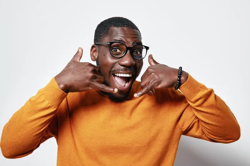African American man with dark skin wearing glasses and orange sweater making call me gesture with hands smiling and looking at camera on white background.