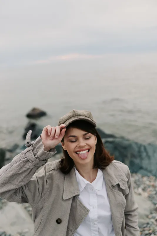 Happy young woman in casual coat and hat enjoys seaside outdoors, smiling and playfully sticking out her tongue. Natural light and rocky beach create relaxed atmosphere.