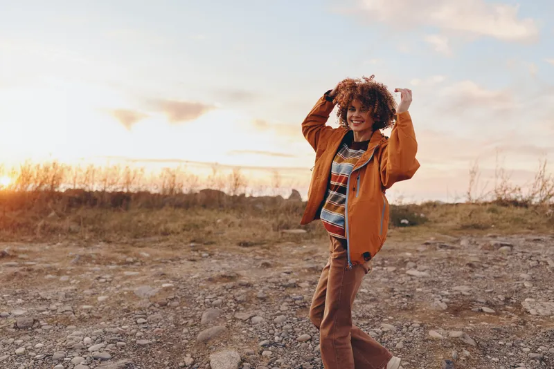 Woman lifestyle outdoors walking at sunset on rocky path, adjusting orange coat and scarf, curly hair lifted by wind, warm golden light, open countryside nature landscape, relaxed smile