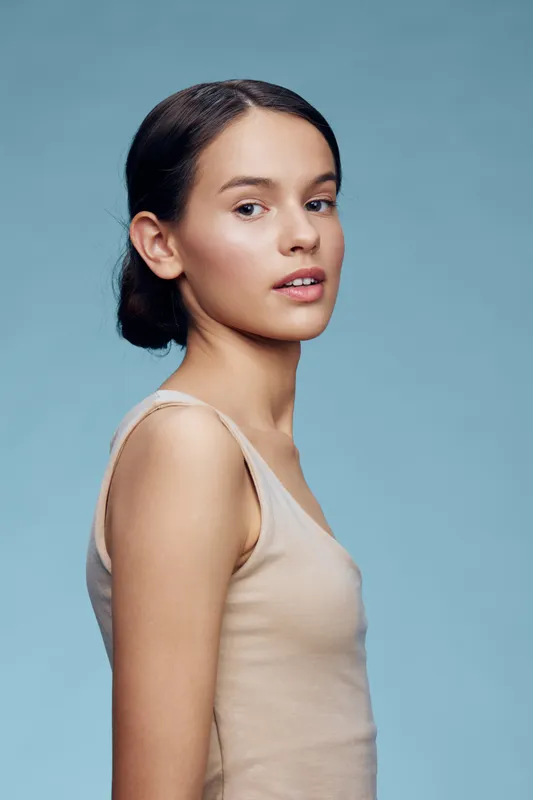 Young woman with a serene expression against a soft blue background, showcasing natural beauty and simplicity in a light beige top
