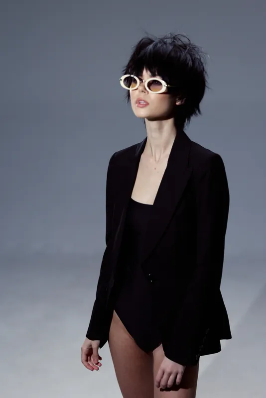 Stylish woman wearing white sunglasses and a black blazer posing against gray background with confident expression.