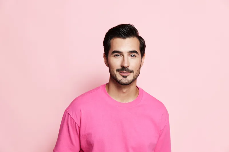 Smiling happy cheerful young man wearing pink sweater looking at camera against plain solid pink background. Joyful expression and bright colors concept.