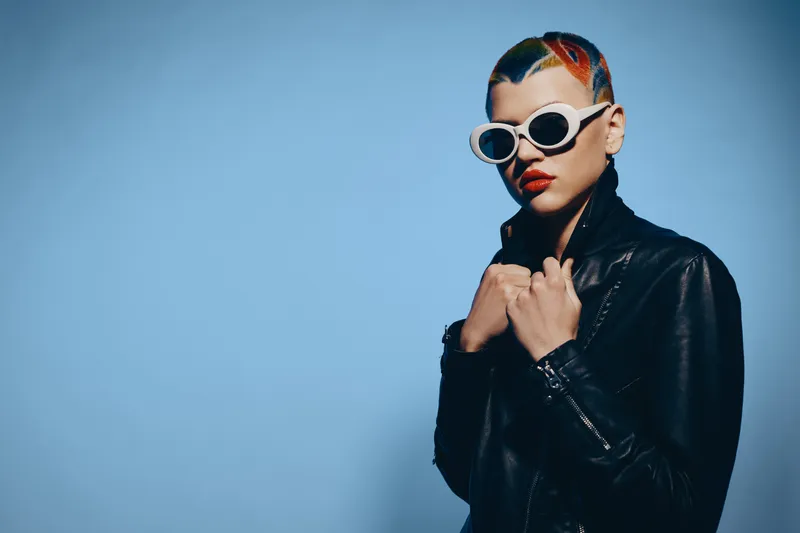 Stylish woman with colorful short hair wearing white sunglasses and a black leather jacket stands confidently against a soft blue background.