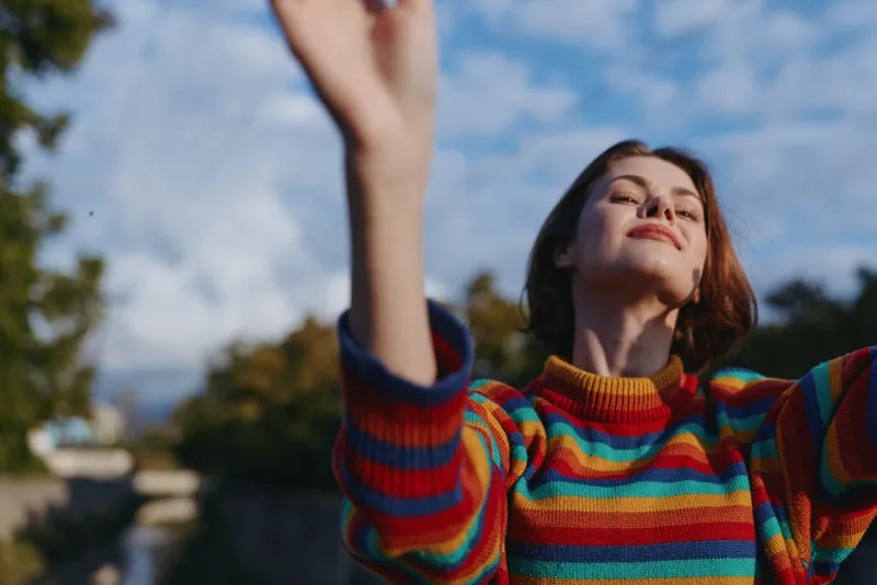 Young woman, striped sweater, smiling outdoors with arms raised, short brown hair under blue sky in park. 20s casual outfit, carefree travel lifestyle, stretching and enjoying warm sunlight.