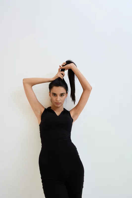young woman in black outfit tying hair against white background, fitness style, minimalist composition, smooth skin, casual pose and natural look