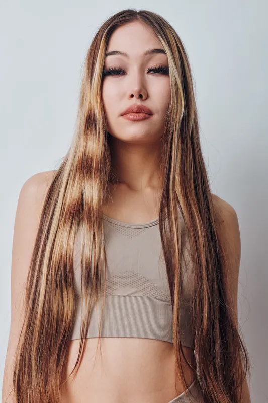 Young woman with long straight hair and neutral makeup posing in light top with crossed arms on soft gray background. Beauty, fashion, and healthy hair concept with confident expression.