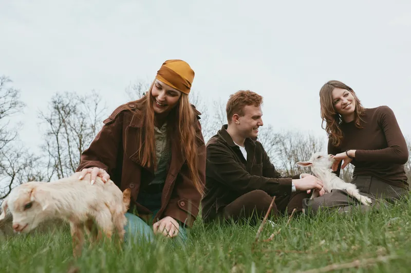 Vibrant outdoor lifestyle with three friends and a curious goat kid on a grassy hillside, casual outfits, candid moment, gentle interaction, natural light, rustic charm, relaxed mood, sense of