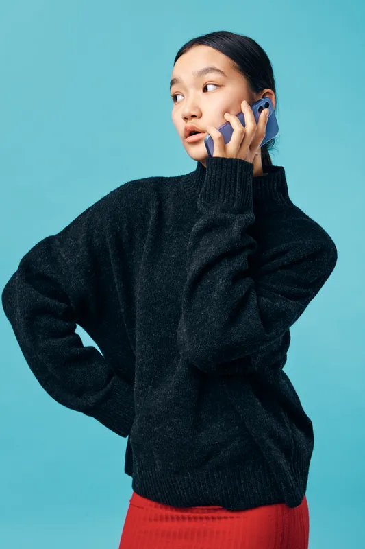 Young woman in black sweater and red skirt talking on smartphone with serious expression on blue background. Communication, technology and lifestyle of female person with casual style.