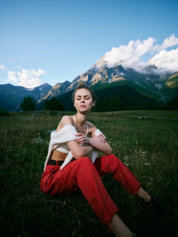 Woman sitting in a grassy meadow with mountains in the background, wearing red pants and a white knit sweater, a peaceful outdoor scene under a blue sky