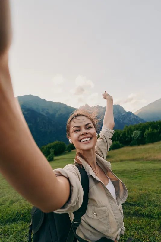 A smiling woman in casual clothes takes a selfie outdoors in a green field with mountains in the background, enjoying a sunny day and nature lifestyle.