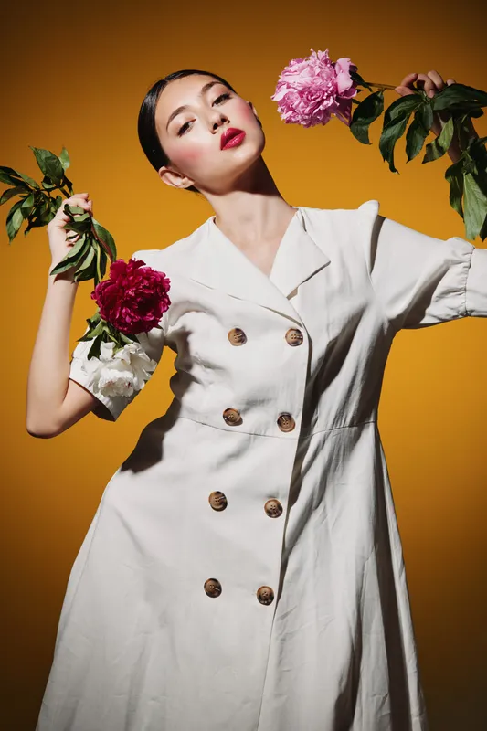 Young elegant woman in a white buttoned dress poses stylishly with colorful flowers against an orange backdrop. Creative beauty and fashion concept with vibrant contrast and expression.