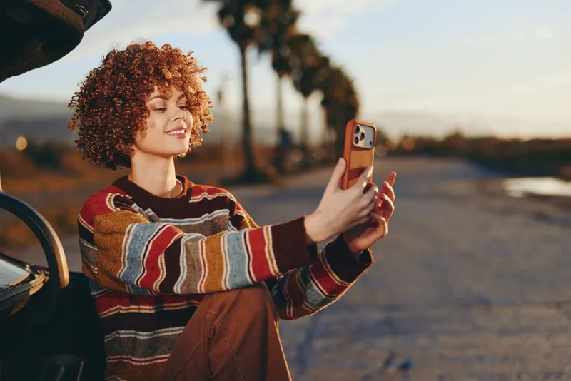 Woman lifestyle outdoors at sunset by the beach, smiling while holding a phone for a selfie beside a bicycle on a promenade, warm golden light, curly hair.