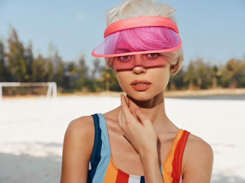Main focus on a fashion-forward model wearing a bright striped swimsuit and pink visor, standing on a sunlit beach court with a soft, cinematic depth of field and relaxed pose.