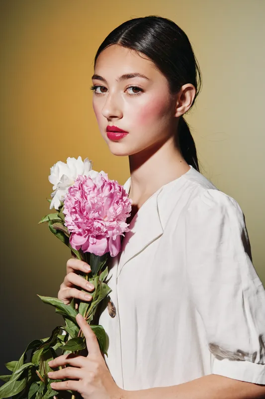 Fashion model with long dark hair holds pink and white peonies wearing a white shirt against a gradient warm background. Stylish beauty portrait, makeup highlight, creative fashion shot.