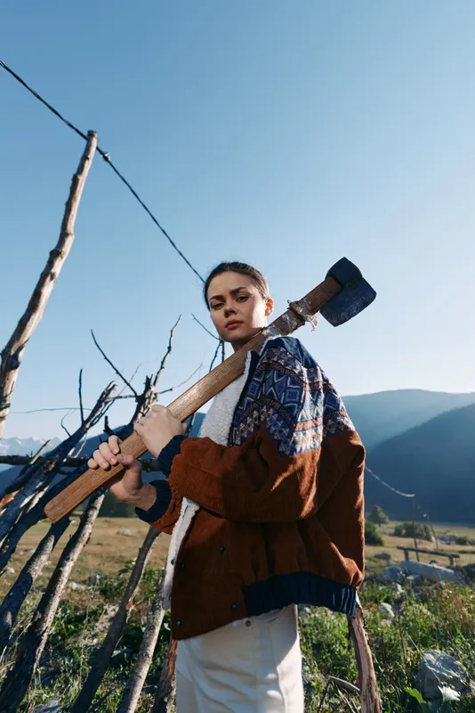 Woman with axe over shoulder by wooden fence in rural mountains landscape, outdoor portrait of a strong farmer or gardener in warm sweater, countryside scene with clear sky.