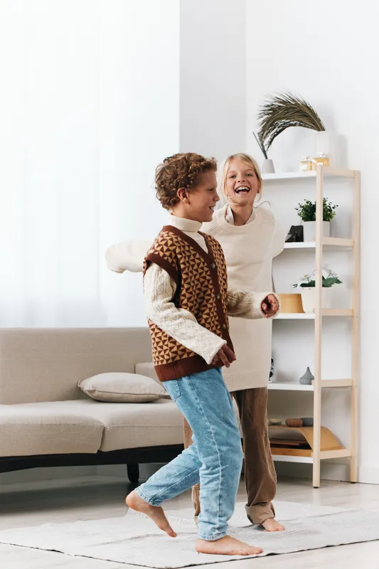 Two cheerful children dance indoors in a bright living room, wearing cozy sweaters and jeans, barefoot on a soft rug, capturing playful energy, warmth, and a carefree home moment.