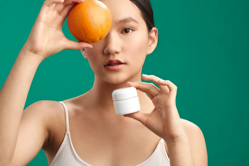 asian woman poses with skincare items in a simple studio setting. she holds an orange near her eye and balances a small jar, presenting a fresh, minimal, beauty oriented mood with soft lighting and