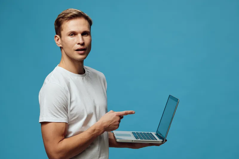 Smiling young man with short hair in casual white t-shirt holding a silver laptop, pointing at screen, standing against plain blue background, studio portrait, people lifestyle concept.