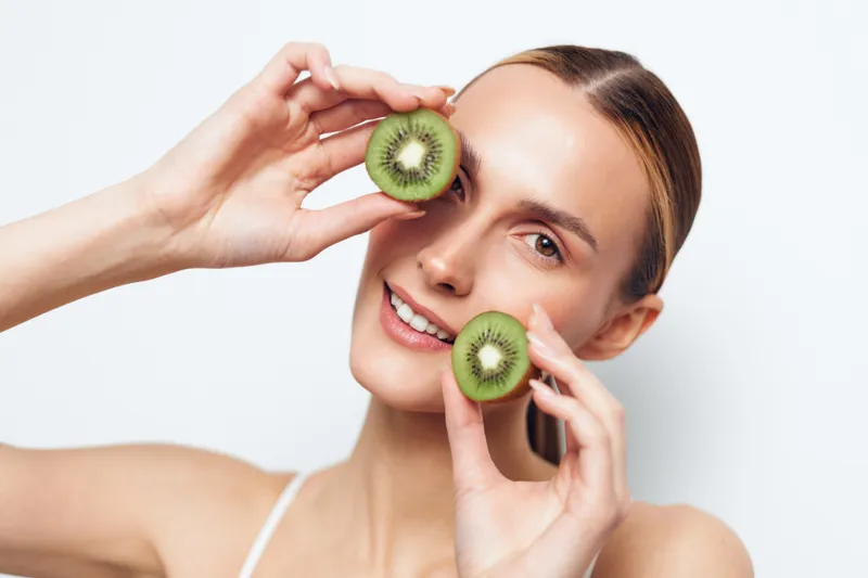 A smiling young woman with clear skin holds two halves of a kiwi fruit near her face against a white background. Concept for healthy lifestyle, natural beauty, and fresh fruits.