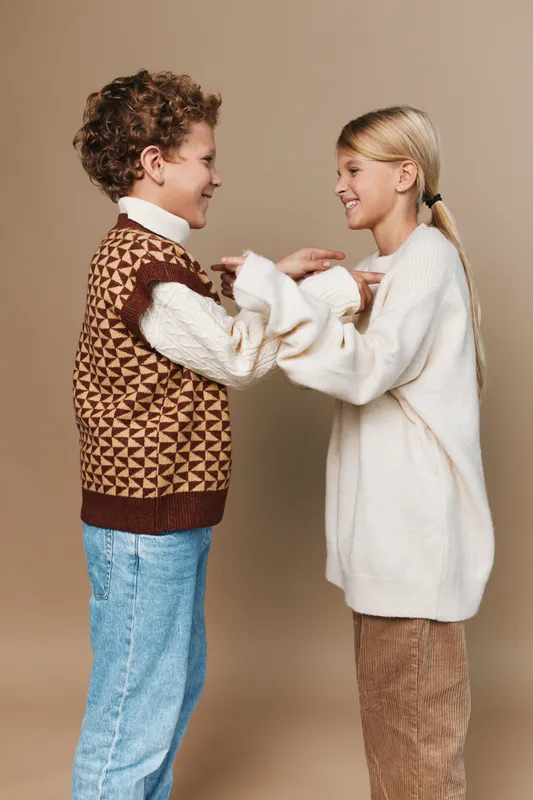Two children share a joyful moment, facing each other with smiles, holding hands in a warm studio setting, wearing cozy sweaters and casual denim for a friendly, timeless portrait.