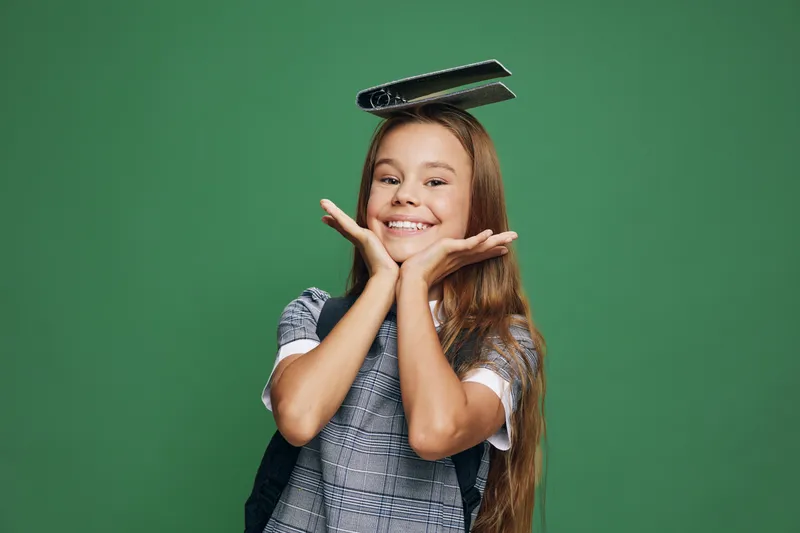 Girl with long hair smiling and holding books on head against green background. Happy child with backpack and casual clothes expressing joy and playfulness in studio.