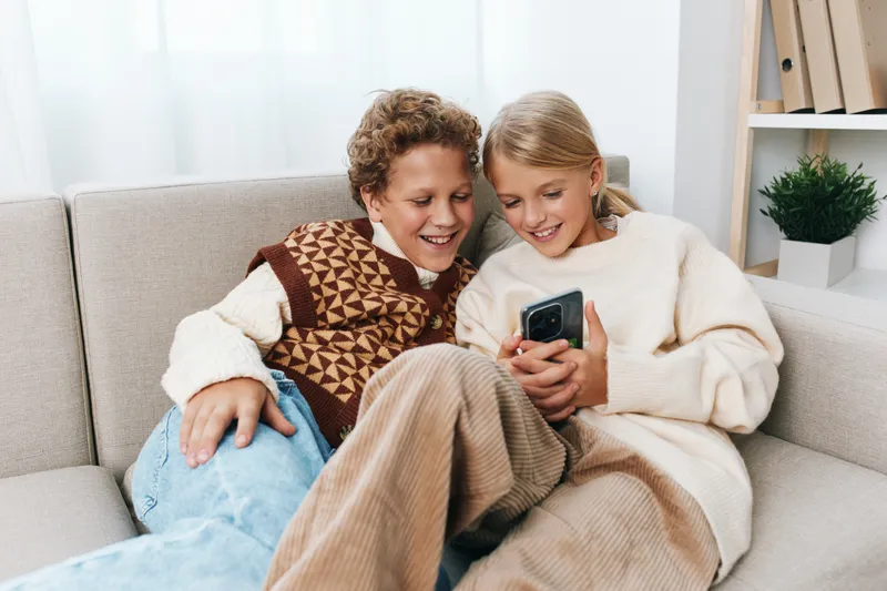 Two friends sit close on a sofa, smiling at a smartphone, sharing a warm, cozy moment in a bright living room with soft light and casual winter attire.